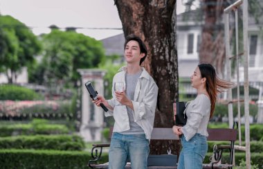 Asyalı çift, parktaki bankta eşine bir buket kırmızı çiçek ve hediye paketi sunan adamla samimi bir anın tadını çıkarıyor. Sıcak, açık hava manzarası aşkı, takdiri ve romantik kutlamayı yansıtıyor.