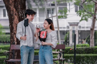 Asyalı çift, parktaki bankta eşine bir buket kırmızı çiçek ve hediye paketi sunan adamla samimi bir anın tadını çıkarıyor. Sıcak, açık hava manzarası aşkı, takdiri ve romantik kutlamayı yansıtıyor.