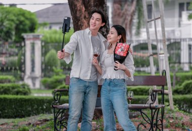 Asyalı çift, parktaki bankta eşine bir buket kırmızı çiçek ve hediye paketi sunan adamla samimi bir anın tadını çıkarıyor. Sıcak, açık hava manzarası aşkı, takdiri ve romantik kutlamayı yansıtıyor.