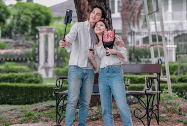 Asyalı çift, parktaki bankta eşine bir buket kırmızı çiçek ve hediye paketi sunan adamla samimi bir anın tadını çıkarıyor. Sıcak, açık hava manzarası aşkı, takdiri ve romantik kutlamayı yansıtıyor.