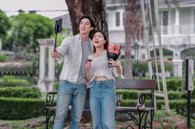 Asyalı çift, parktaki bankta eşine bir buket kırmızı çiçek ve hediye paketi sunan adamla samimi bir anın tadını çıkarıyor. Sıcak, açık hava manzarası aşkı, takdiri ve romantik kutlamayı yansıtıyor.