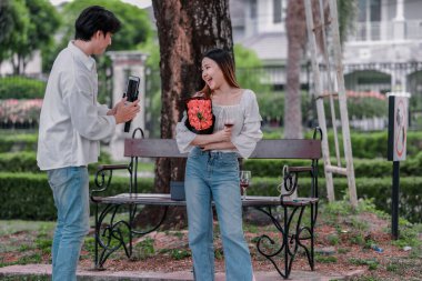 Asyalı çift, parktaki bankta eşine bir buket kırmızı çiçek ve hediye paketi sunan adamla samimi bir anın tadını çıkarıyor. Sıcak, açık hava manzarası aşkı, takdiri ve romantik kutlamayı yansıtıyor.