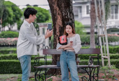 Asyalı çift, parktaki bankta eşine bir buket kırmızı çiçek ve hediye paketi sunan adamla samimi bir anın tadını çıkarıyor. Sıcak, açık hava manzarası aşkı, takdiri ve romantik kutlamayı yansıtıyor.