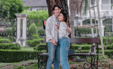 Asyalı çift, parktaki bankta eşine bir buket kırmızı çiçek ve hediye paketi sunan adamla samimi bir anın tadını çıkarıyor. Sıcak, açık hava manzarası aşkı, takdiri ve romantik kutlamayı yansıtıyor.