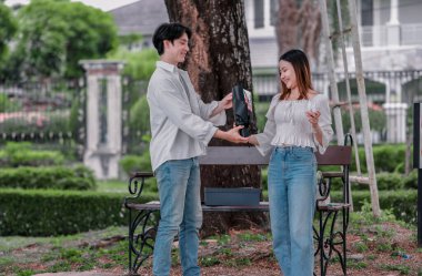 Asyalı çift, parktaki bankta eşine bir buket kırmızı çiçek ve hediye paketi sunan adamla samimi bir anın tadını çıkarıyor. Sıcak, açık hava manzarası aşkı, takdiri ve romantik kutlamayı yansıtıyor.