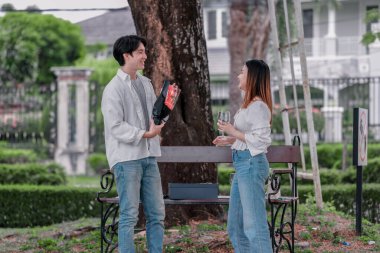 Asyalı çift, parktaki bankta eşine bir buket kırmızı çiçek ve hediye paketi sunan adamla samimi bir anın tadını çıkarıyor. Sıcak, açık hava manzarası aşkı, takdiri ve romantik kutlamayı yansıtıyor.