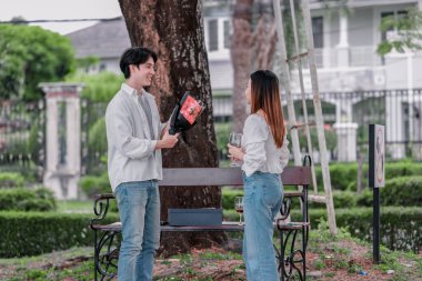 Asyalı çift, parktaki bankta eşine bir buket kırmızı çiçek ve hediye paketi sunan adamla samimi bir anın tadını çıkarıyor. Sıcak, açık hava manzarası aşkı, takdiri ve romantik kutlamayı yansıtıyor.