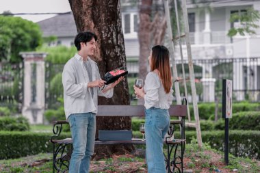 Asyalı çift, parktaki bankta eşine bir buket kırmızı çiçek ve hediye paketi sunan adamla samimi bir anın tadını çıkarıyor. Sıcak, açık hava manzarası aşkı, takdiri ve romantik kutlamayı yansıtıyor.