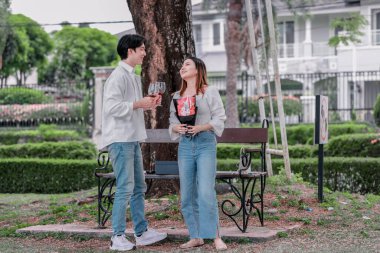 Asyalı çift, parktaki bankta eşine bir buket kırmızı çiçek ve hediye paketi sunan adamla samimi bir anın tadını çıkarıyor. Sıcak, açık hava manzarası aşkı, takdiri ve romantik kutlamayı yansıtıyor.