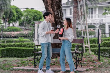 Asyalı çift, parktaki bankta eşine bir buket kırmızı çiçek ve hediye paketi sunan adamla samimi bir anın tadını çıkarıyor. Sıcak, açık hava manzarası aşkı, takdiri ve romantik kutlamayı yansıtıyor.