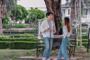 Asyalı çift, parktaki bankta eşine bir buket kırmızı çiçek ve hediye paketi sunan adamla samimi bir anın tadını çıkarıyor. Sıcak, açık hava manzarası aşkı, takdiri ve romantik kutlamayı yansıtıyor.