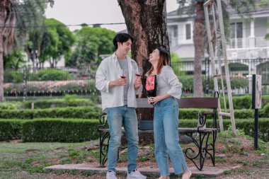 Asyalı çift, parktaki bankta eşine bir buket kırmızı çiçek ve hediye paketi sunan adamla samimi bir anın tadını çıkarıyor. Sıcak, açık hava manzarası aşkı, takdiri ve romantik kutlamayı yansıtıyor.