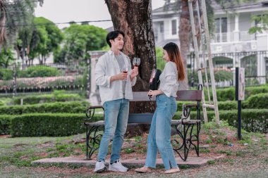 Asyalı çift, parktaki bankta eşine bir buket kırmızı çiçek ve hediye paketi sunan adamla samimi bir anın tadını çıkarıyor. Sıcak, açık hava manzarası aşkı, takdiri ve romantik kutlamayı yansıtıyor.