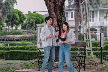 Asyalı çift, parktaki bankta eşine bir buket kırmızı çiçek ve hediye paketi sunan adamla samimi bir anın tadını çıkarıyor. Sıcak, açık hava manzarası aşkı, takdiri ve romantik kutlamayı yansıtıyor.