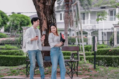 Asyalı çift, parktaki bankta eşine bir buket kırmızı çiçek ve hediye paketi sunan adamla samimi bir anın tadını çıkarıyor. Sıcak, açık hava manzarası aşkı, takdiri ve romantik kutlamayı yansıtıyor.