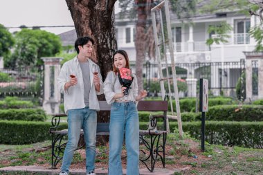 Asyalı çift, parktaki bankta eşine bir buket kırmızı çiçek ve hediye paketi sunan adamla samimi bir anın tadını çıkarıyor. Sıcak, açık hava manzarası aşkı, takdiri ve romantik kutlamayı yansıtıyor.