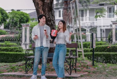 Asyalı çift, parktaki bankta eşine bir buket kırmızı çiçek ve hediye paketi sunan adamla samimi bir anın tadını çıkarıyor. Sıcak, açık hava manzarası aşkı, takdiri ve romantik kutlamayı yansıtıyor.