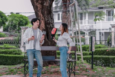 Asyalı çift, parktaki bankta eşine bir buket kırmızı çiçek ve hediye paketi sunan adamla samimi bir anın tadını çıkarıyor. Sıcak, açık hava manzarası aşkı, takdiri ve romantik kutlamayı yansıtıyor.