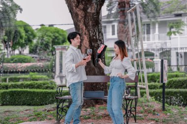 Asyalı çift, parktaki bankta eşine bir buket kırmızı çiçek ve hediye paketi sunan adamla samimi bir anın tadını çıkarıyor. Sıcak, açık hava manzarası aşkı, takdiri ve romantik kutlamayı yansıtıyor.