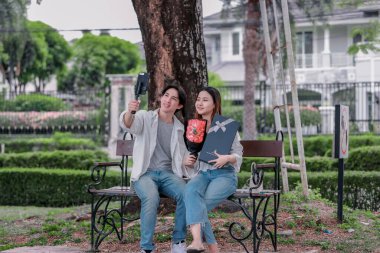 Asyalı çift, parktaki bankta eşine bir buket kırmızı çiçek ve hediye paketi sunan adamla samimi bir anın tadını çıkarıyor. Sıcak, açık hava manzarası aşkı, takdiri ve romantik kutlamayı yansıtıyor.