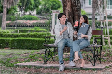 Asyalı çift, parktaki bankta eşine bir buket kırmızı çiçek ve hediye paketi sunan adamla samimi bir anın tadını çıkarıyor. Sıcak, açık hava manzarası aşkı, takdiri ve romantik kutlamayı yansıtıyor.