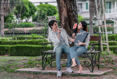 Asyalı çift, parktaki bankta eşine bir buket kırmızı çiçek ve hediye paketi sunan adamla samimi bir anın tadını çıkarıyor. Sıcak, açık hava manzarası aşkı, takdiri ve romantik kutlamayı yansıtıyor.