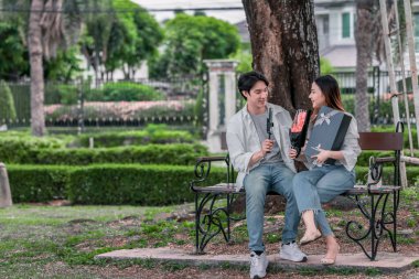 Asyalı çift, parktaki bankta eşine bir buket kırmızı çiçek ve hediye paketi sunan adamla samimi bir anın tadını çıkarıyor. Sıcak, açık hava manzarası aşkı, takdiri ve romantik kutlamayı yansıtıyor.