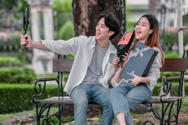 Asyalı çift, parktaki bankta eşine bir buket kırmızı çiçek ve hediye paketi sunan adamla samimi bir anın tadını çıkarıyor. Sıcak, açık hava manzarası aşkı, takdiri ve romantik kutlamayı yansıtıyor.