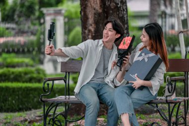 Asyalı çift, parktaki bankta eşine bir buket kırmızı çiçek ve hediye paketi sunan adamla samimi bir anın tadını çıkarıyor. Sıcak, açık hava manzarası aşkı, takdiri ve romantik kutlamayı yansıtıyor.