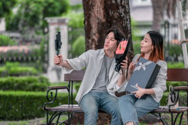 Asyalı çift, parktaki bankta eşine bir buket kırmızı çiçek ve hediye paketi sunan adamla samimi bir anın tadını çıkarıyor. Sıcak, açık hava manzarası aşkı, takdiri ve romantik kutlamayı yansıtıyor.