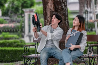 Asyalı çift, parktaki bankta eşine bir buket kırmızı çiçek ve hediye paketi sunan adamla samimi bir anın tadını çıkarıyor. Sıcak, açık hava manzarası aşkı, takdiri ve romantik kutlamayı yansıtıyor.