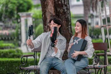 Asyalı çift, parktaki bankta eşine bir buket kırmızı çiçek ve hediye paketi sunan adamla samimi bir anın tadını çıkarıyor. Sıcak, açık hava manzarası aşkı, takdiri ve romantik kutlamayı yansıtıyor.