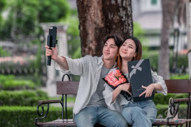 Asyalı çift, parktaki bankta eşine bir buket kırmızı çiçek ve hediye paketi sunan adamla samimi bir anın tadını çıkarıyor. Sıcak, açık hava manzarası aşkı, takdiri ve romantik kutlamayı yansıtıyor.