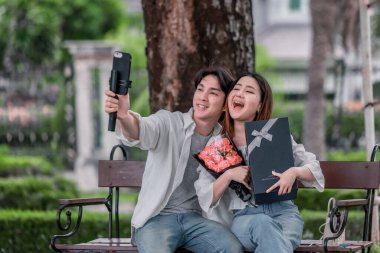 Asyalı çift, parktaki bankta eşine bir buket kırmızı çiçek ve hediye paketi sunan adamla samimi bir anın tadını çıkarıyor. Sıcak, açık hava manzarası aşkı, takdiri ve romantik kutlamayı yansıtıyor.