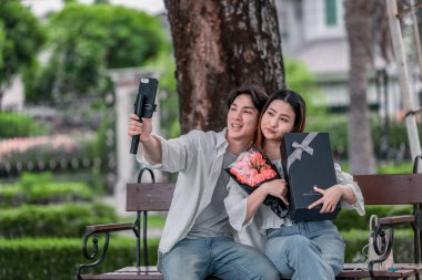 Asyalı çift, parktaki bankta eşine bir buket kırmızı çiçek ve hediye paketi sunan adamla samimi bir anın tadını çıkarıyor. Sıcak, açık hava manzarası aşkı, takdiri ve romantik kutlamayı yansıtıyor.