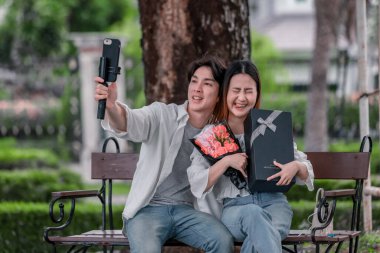 Asyalı çift, parktaki bankta eşine bir buket kırmızı çiçek ve hediye paketi sunan adamla samimi bir anın tadını çıkarıyor. Sıcak, açık hava manzarası aşkı, takdiri ve romantik kutlamayı yansıtıyor.