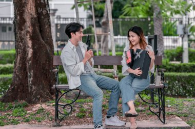 Asyalı çift, parktaki bankta eşine bir buket kırmızı çiçek ve hediye paketi sunan adamla samimi bir anın tadını çıkarıyor. Sıcak, açık hava manzarası aşkı, takdiri ve romantik kutlamayı yansıtıyor.