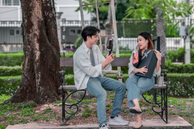 Asyalı çift, parktaki bankta eşine bir buket kırmızı çiçek ve hediye paketi sunan adamla samimi bir anın tadını çıkarıyor. Sıcak, açık hava manzarası aşkı, takdiri ve romantik kutlamayı yansıtıyor.