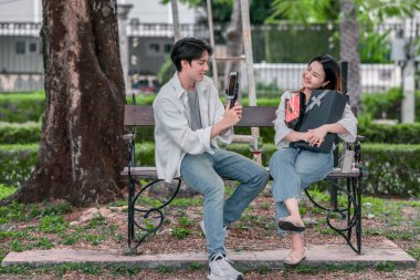 Asyalı çift, parktaki bankta eşine bir buket kırmızı çiçek ve hediye paketi sunan adamla samimi bir anın tadını çıkarıyor. Sıcak, açık hava manzarası aşkı, takdiri ve romantik kutlamayı yansıtıyor.