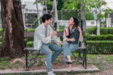 Asyalı çift, parktaki bankta eşine bir buket kırmızı çiçek ve hediye paketi sunan adamla samimi bir anın tadını çıkarıyor. Sıcak, açık hava manzarası aşkı, takdiri ve romantik kutlamayı yansıtıyor.