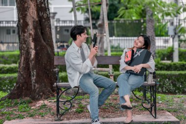 Asyalı çift, parktaki bankta eşine bir buket kırmızı çiçek ve hediye paketi sunan adamla samimi bir anın tadını çıkarıyor. Sıcak, açık hava manzarası aşkı, takdiri ve romantik kutlamayı yansıtıyor.