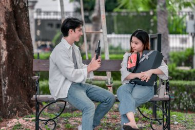 Asyalı çift, parktaki bankta eşine bir buket kırmızı çiçek ve hediye paketi sunan adamla samimi bir anın tadını çıkarıyor. Sıcak, açık hava manzarası aşkı, takdiri ve romantik kutlamayı yansıtıyor.