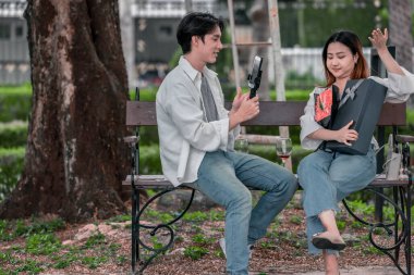 Asyalı çift, parktaki bankta eşine bir buket kırmızı çiçek ve hediye paketi sunan adamla samimi bir anın tadını çıkarıyor. Sıcak, açık hava manzarası aşkı, takdiri ve romantik kutlamayı yansıtıyor.