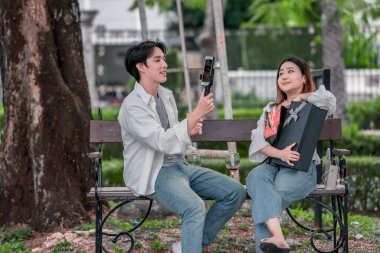 Asyalı çift, parktaki bankta eşine bir buket kırmızı çiçek ve hediye paketi sunan adamla samimi bir anın tadını çıkarıyor. Sıcak, açık hava manzarası aşkı, takdiri ve romantik kutlamayı yansıtıyor.