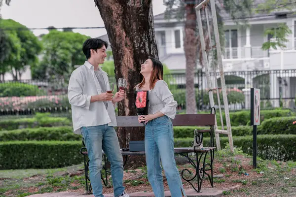 Asyalı çift, parktaki bankta eşine bir buket kırmızı çiçek ve hediye paketi sunan adamla samimi bir anın tadını çıkarıyor. Sıcak, açık hava manzarası aşkı, takdiri ve romantik kutlamayı yansıtıyor.