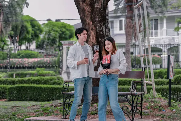 Asyalı çift, parktaki bankta eşine bir buket kırmızı çiçek ve hediye paketi sunan adamla samimi bir anın tadını çıkarıyor. Sıcak, açık hava manzarası aşkı, takdiri ve romantik kutlamayı yansıtıyor.