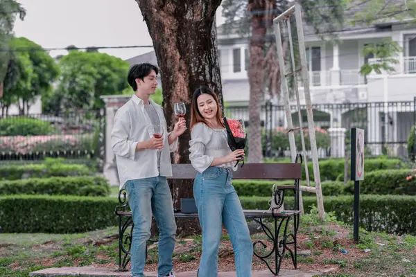 Asyalı çift, parktaki bankta eşine bir buket kırmızı çiçek ve hediye paketi sunan adamla samimi bir anın tadını çıkarıyor. Sıcak, açık hava manzarası aşkı, takdiri ve romantik kutlamayı yansıtıyor.