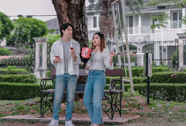 Asyalı çift, parktaki bankta eşine bir buket kırmızı çiçek ve hediye paketi sunan adamla samimi bir anın tadını çıkarıyor. Sıcak, açık hava manzarası aşkı, takdiri ve romantik kutlamayı yansıtıyor.