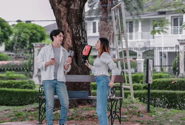 Asyalı çift, parktaki bankta eşine bir buket kırmızı çiçek ve hediye paketi sunan adamla samimi bir anın tadını çıkarıyor. Sıcak, açık hava manzarası aşkı, takdiri ve romantik kutlamayı yansıtıyor.