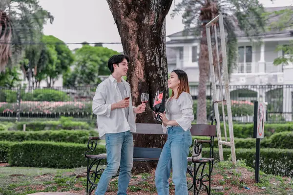 Asyalı çift, parktaki bankta eşine bir buket kırmızı çiçek ve hediye paketi sunan adamla samimi bir anın tadını çıkarıyor. Sıcak, açık hava manzarası aşkı, takdiri ve romantik kutlamayı yansıtıyor.